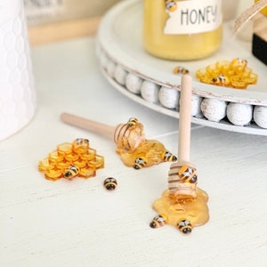 Può includere: Un mestolo di legno per il miele con un motivo a nido d'ape giallo e una piccola figurina di ape in cima. Il mestolo è circondato da una pozza di miele e da diverse piccole figurine di api. Il mestolo è su una superficie bianca con un piccolo barattolo di miele sullo sfondo.