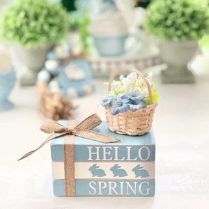 Può includere: Una pila di tre blocchi di legno dipinti di blu con le parole "Hello Spring" e tre sagome di conigli. Un piccolo cestino di vimini pieno di fiori bianchi e blu è posto sopra i blocchi.