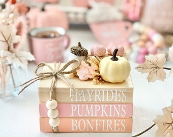May include: A stack of three wooden books with pink and orange painted covers. The books are decorated with a small white pumpkin, fall leaves, and a brown twine bow. The books are labeled "Hayrides", "Pumpkins", and "Bonfires".