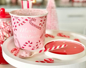 Puede incluir: Un vaso de papel rosa y rojo con un patrón de bastón de caramelo y el texto "Merry Christmas". El vaso está lleno de un postre rosa y blanco. Una cuchara rayada roja y blanca está en un soporte de pastel blanco con un bastón de caramelo rojo y blanco en el lateral.
