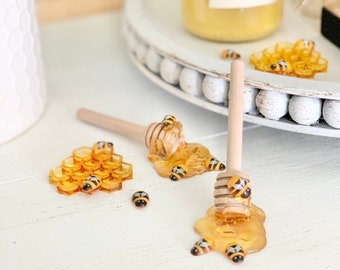Puede incluir: Un cucharón de miel de madera con un diseño de panal de abeja amarillo y una pequeña figura de abeja en la parte superior. El cucharón está rodeado de un charco de miel y varias pequeñas figuras de abejas. El cucharón está sobre una superficie blanca con un pequeño tarro de miel en el fondo.