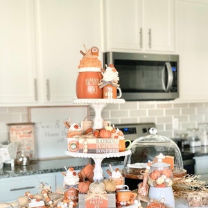 May include: A three-tiered white tiered tray decorated with fall-themed items, including pumpkins, gourds, cinnamon sticks, and mugs. The tray is decorated with the words "Hayrides, Pumpkins, Bonfires" and "Pumpkin Patch 25".