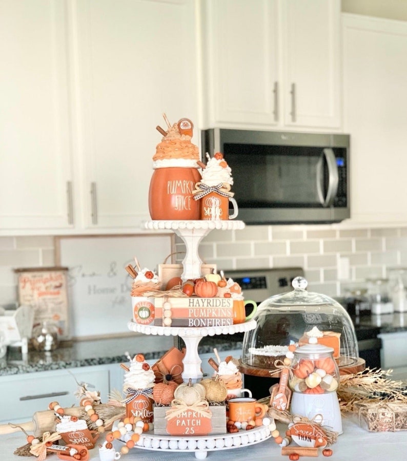 May include: A three-tiered white tiered tray decorated with fall-themed items, including pumpkins, gourds, cinnamon sticks, and mugs. The tray is decorated with the words "Hayrides, Pumpkins, Bonfires" and "Pumpkin Patch 25".