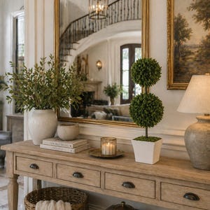 May include: A light wood console table with four drawers and a lower shelf, featuring a white vase with green foliage, a double-ball topiary in a white square pot, a glass candle holder, a woven basket with a cloth, and stacked books.