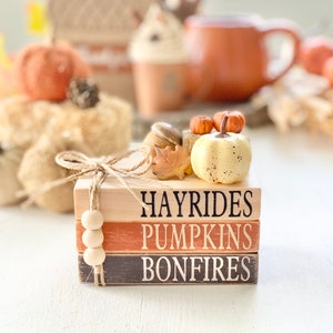 May include: A stack of three wooden blocks with the words "Hayrides", "Pumpkins", and "Bonfires" painted on them. The blocks are decorated with a small white pumpkin, two small orange pumpkins, a brown acorn, and a brown leaf.