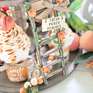 May include: A tiered wooden tray with peach-themed decorations. A small wooden ladder with a sign that reads "FRESH PICKED PEACHES" is adorned with peach and leaf accents. Jars of peach preserves and whipped cream desserts are also visible.