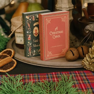 May include: Two vintage-style books, one red and one dark green, sit on a wooden tray. The red book reads "A Christmas Carol." The green book has illustrations and text. The scene includes dried oranges, cinnamon sticks, and greenery.