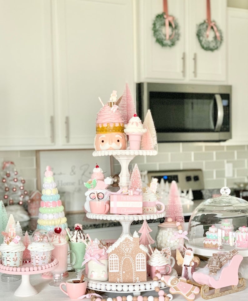Puede incluir: Un soporte blanco de tres niveles decorado con adornos navide&ntilde;os rosas y blancos. El soporte presenta una taza de Pap&aacute; Noel rosa y dorada, una taza de mu&ntilde;eco de nieve rosa y blanca, una casa de jengibre y un trineo rosa y blanco. El soporte est&aacute; rodeado de otras decoraciones navide&ntilde;as, incluyendo un &aacute;rbol de Navidad rosa y blanco, un bast&oacute;n de caramelo rosa y blanco y una magdalena rosa y blanca.