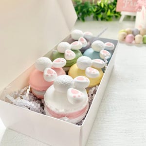 May include: A white box filled with colorful macarons, each decorated with a bunny theme. The macarons are in pastel colors like pink, green, yellow, and blue, with white and pink accents. The box is lined with white paper shreds.