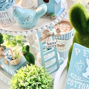 May include: A tiered tray with Easter-themed decorations. Includes a blue bird figurine, a miniature birdhouse, a small ladder with a "Happy Easter" sign, and a green bunny figure. Other items include a mug with "Happy Easter" text and a sign that says "Cotton Farm".