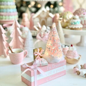 Puede incluir: Una pila de bloques de madera rosa y blanca con el texto "I'm Dreaming of a Pink Christmas" en el bloque inferior. Los bloques est&aacute;n decorados con una cinta rosa y un peque&ntilde;o &aacute;rbol de Navidad blanco con luces. Tambi&eacute;n hay dos peque&ntilde;as cajas de regalo en la parte superior de los bloques.
