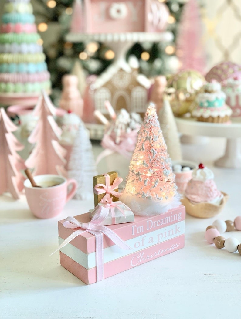 Puede incluir: Una pila de bloques de madera rosa y blanca con el texto "I'm Dreaming of a Pink Christmas" en el bloque inferior. Los bloques est&aacute;n decorados con una cinta rosa y un peque&ntilde;o &aacute;rbol de Navidad blanco con luces. Tambi&eacute;n hay dos peque&ntilde;as cajas de regalo en la parte superior de los bloques.