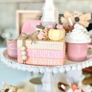 Puede incluir: Una bandeja de niveles decorada con art&iacute;culos de tem&aacute;tica oto&ntilde;al, que incluyen una pila de tres bloques de madera con las palabras "Hayrides", "Pumpkins" y "Bonfires" en letras blancas, una peque&ntilde;a calabaza de cer&aacute;mica blanca y una taza rosa con crema batida.