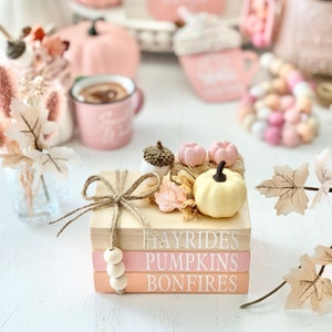 Puede incluir: Una pila de tres bloques de madera con las palabras "Hayrides", "Pumpkins" y "Bonfires" pintadas en blanco. Los bloques est&aacute;n decorados con una peque&ntilde;a calabaza, hojas de oto&ntilde;o y un lazo de cuerda.