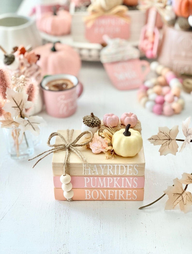 Puede incluir: Una pila de tres bloques de madera con las palabras "Hayrides", "Pumpkins" y "Bonfires" pintadas en blanco. Los bloques est&aacute;n decorados con una peque&ntilde;a calabaza, hojas de oto&ntilde;o y un lazo de cuerda.