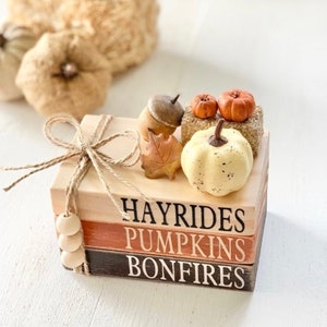 May include: A stack of three wooden blocks with the words "Hayrides", "Pumpkins", and "Bonfires" painted on them. The blocks are decorated with a small white pumpkin, two orange pumpkins, an acorn, and a fall leaf.