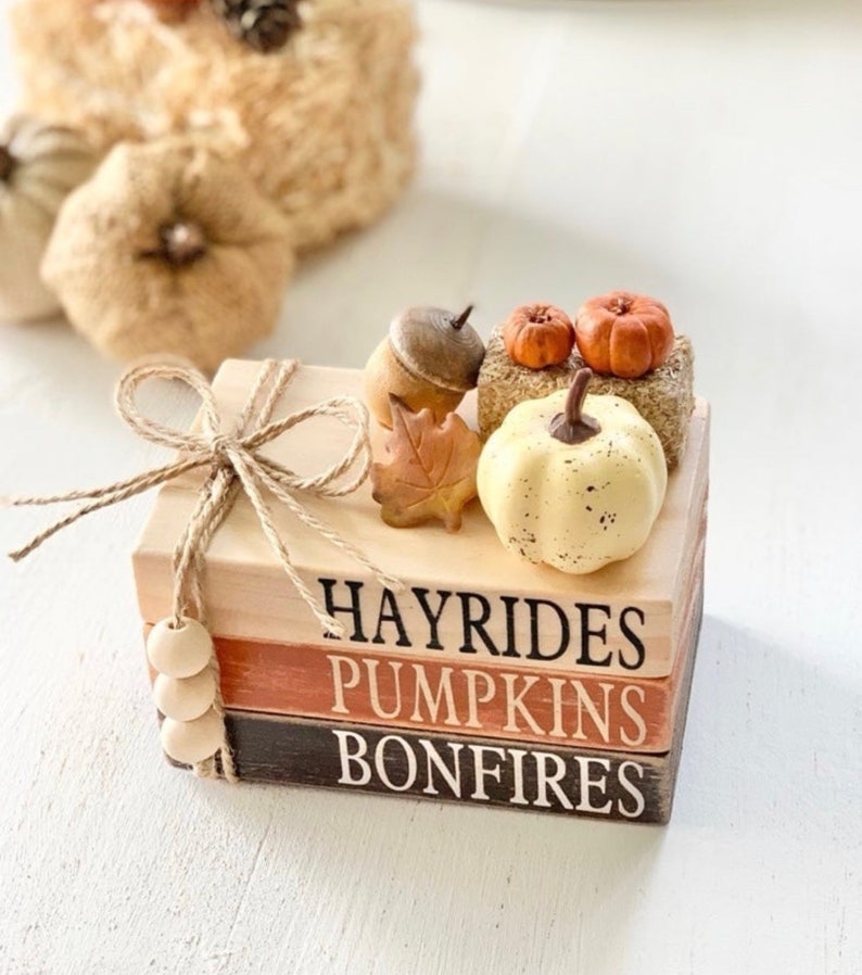 May include: A stack of three wooden blocks with the words "Hayrides", "Pumpkins", and "Bonfires" painted on them. The blocks are decorated with a small white pumpkin, two orange pumpkins, an acorn, and a fall leaf.