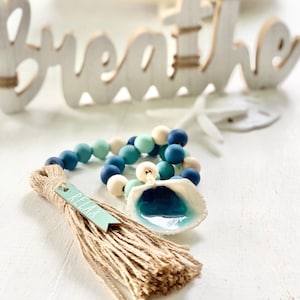 Puede incluir: Una guirnalda de cuentas azules y verdes con un dije de concha y un borla con una etiqueta que dice "Relax". La guirnalda está colgada de un letrero de madera que dice "Breathe".