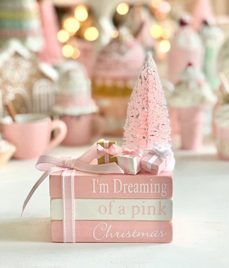 Puede incluir: Una pila de tres bloques de madera pintados de rosa con letras blancas. El bloque superior dice "I'm Dreaming of a pink Christmas". Los bloques est&aacute;n decorados con una cinta rosa y un peque&ntilde;o &aacute;rbol de Navidad rosa con purpurina blanca. Tambi&eacute;n hay peque&ntilde;os regalos envueltos en la parte superior de los bloques.