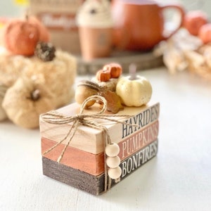 May include: A stack of three wooden blocks with the words "Hayrides", "Pumpkins", and "Bonfires" painted on them. The blocks are decorated with a small pumpkin, leaves, and twine.