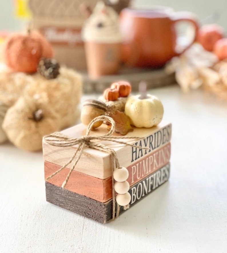 May include: A stack of three wooden blocks with the words "Hayrides", "Pumpkins", and "Bonfires" painted on them. The blocks are decorated with a small pumpkin, leaves, and twine.