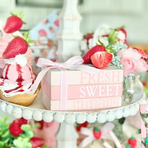 Puede incluir: Un soporte para pasteles blanco de varios niveles con un juego de libros apilados de color rosa y blanco que dice "Fresh Sweet Strawberries". El soporte está decorado con fresas falsas, flores y otros elementos decorativos.