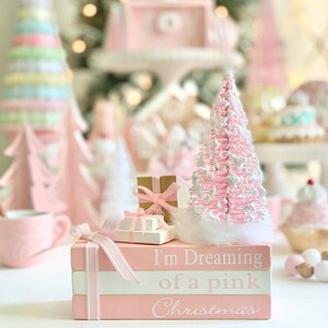 Puede incluir: Una pila de tres libros rosas con letras blancas que dicen "I'm Dreaming of a pink Christmas". Un peque&ntilde;o &aacute;rbol de Navidad rosa y blanco se sienta encima de los libros con una peque&ntilde;a caja de regalo en frente.