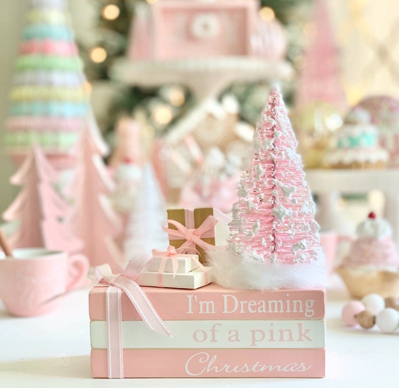 Puede incluir: Una pila de tres libros rosas con letras blancas que dicen "I'm Dreaming of a pink Christmas". Un peque&ntilde;o &aacute;rbol de Navidad rosa y blanco se sienta encima de los libros con una peque&ntilde;a caja de regalo en frente.
