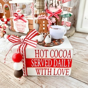 Op de afbeelding: Een wit houten blokschild met rood-wit gestreept lint en rode kralen. Het schild heeft de tekst "Hot Cocoa Served Daily With Love". Een klein wit kopje met bruine vloeistof en witte marshmallows staat op het schild.