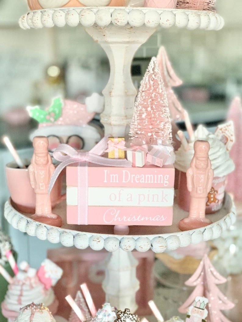 Puede incluir: Una bandeja de tres niveles decorada con adornos navide&ntilde;os rosas y blancos. El nivel superior presenta un &aacute;rbol de Navidad rosa, una pila de cajas de regalo rosas y blancas y un letrero de madera que dice "Estoy so&ntilde;ando con una Navidad rosa".