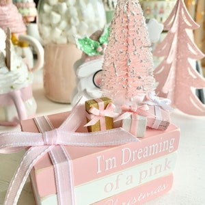 Puede incluir: Una pila de tres libros rosas y blancos con una cinta rosa atada alrededor. Los libros est&aacute;n decorados con las palabras "I'm Dreaming of a Pink Christmas". Hay dos peque&ntilde;as cajas de regalo en la parte superior de los libros.