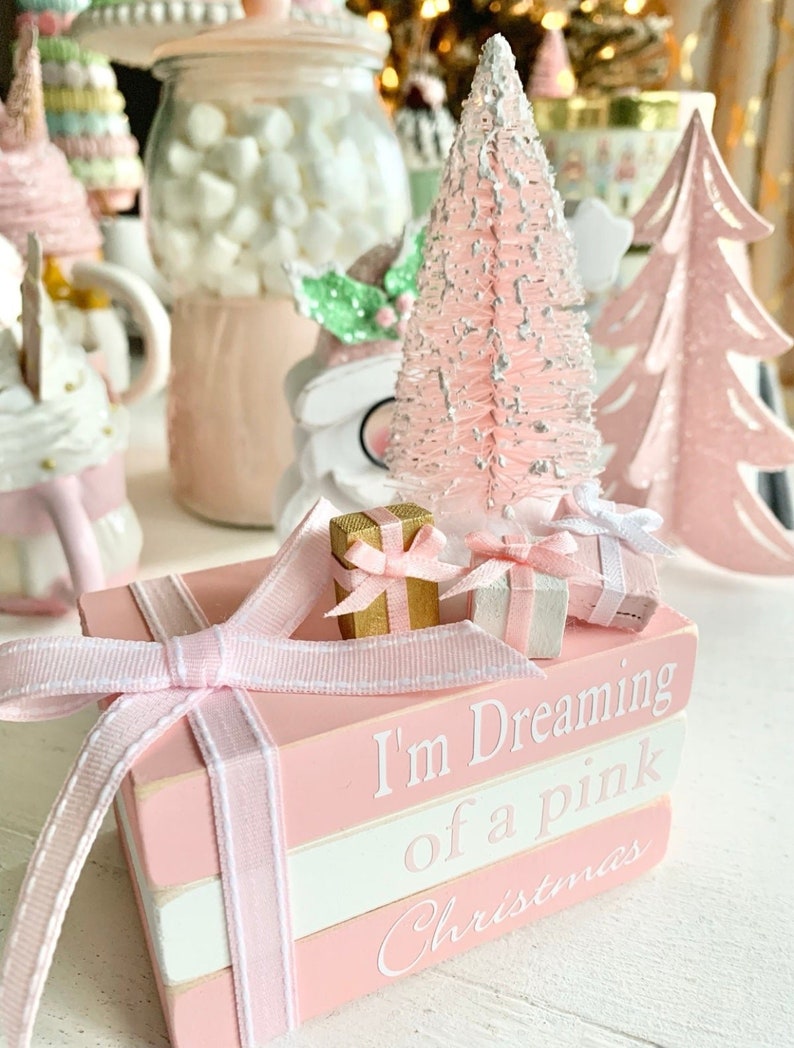 Puede incluir: Una pila de tres libros rosas y blancos con una cinta rosa atada alrededor. Los libros est&aacute;n decorados con las palabras "I'm Dreaming of a Pink Christmas". Hay dos peque&ntilde;as cajas de regalo en la parte superior de los libros.