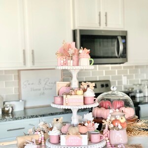 Puede incluir: Una bandeja de tres niveles blanca decorada con art&iacute;culos de tem&aacute;tica oto&ntilde;al en tonos rosa y blanco. La bandeja presenta calabazas en miniatura, hojas de oto&ntilde;o, una casita y un letrero que dice "Pumpkin Patch".