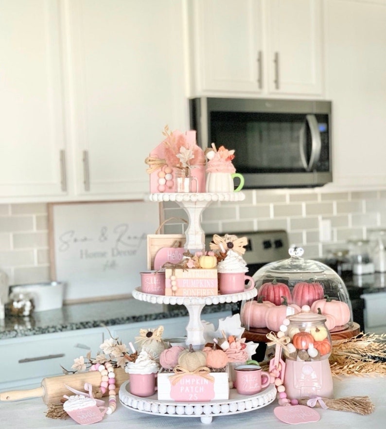 Puede incluir: Una bandeja de tres niveles blanca decorada con art&iacute;culos de tem&aacute;tica oto&ntilde;al en tonos rosa y blanco. La bandeja presenta calabazas en miniatura, hojas de oto&ntilde;o, una casita y un letrero que dice "Pumpkin Patch".
