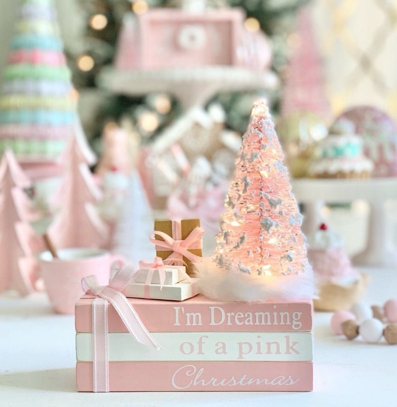 Puede incluir: Una pila de tres libros rosas con detalles blancos y una cinta rosa. El libro de arriba dice "I'm Dreaming of a Pink Christmas". Un peque&ntilde;o &aacute;rbol de Navidad rosa con luces blancas y una base blanca y esponjosa se encuentra encima de los libros. Tambi&eacute;n hay dos peque&ntilde;os regalos envueltos encima de los libros.