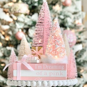 Puede incluir: Un conjunto de mini &aacute;rboles de Navidad rosas y blancos con luces, una caja de regalo rosa y una pila de libros rosas con el texto "I'm Dreaming of a Pink Christmas". Los &aacute;rboles est&aacute;n sobre un pedestal blanco con un borde de cuentas.