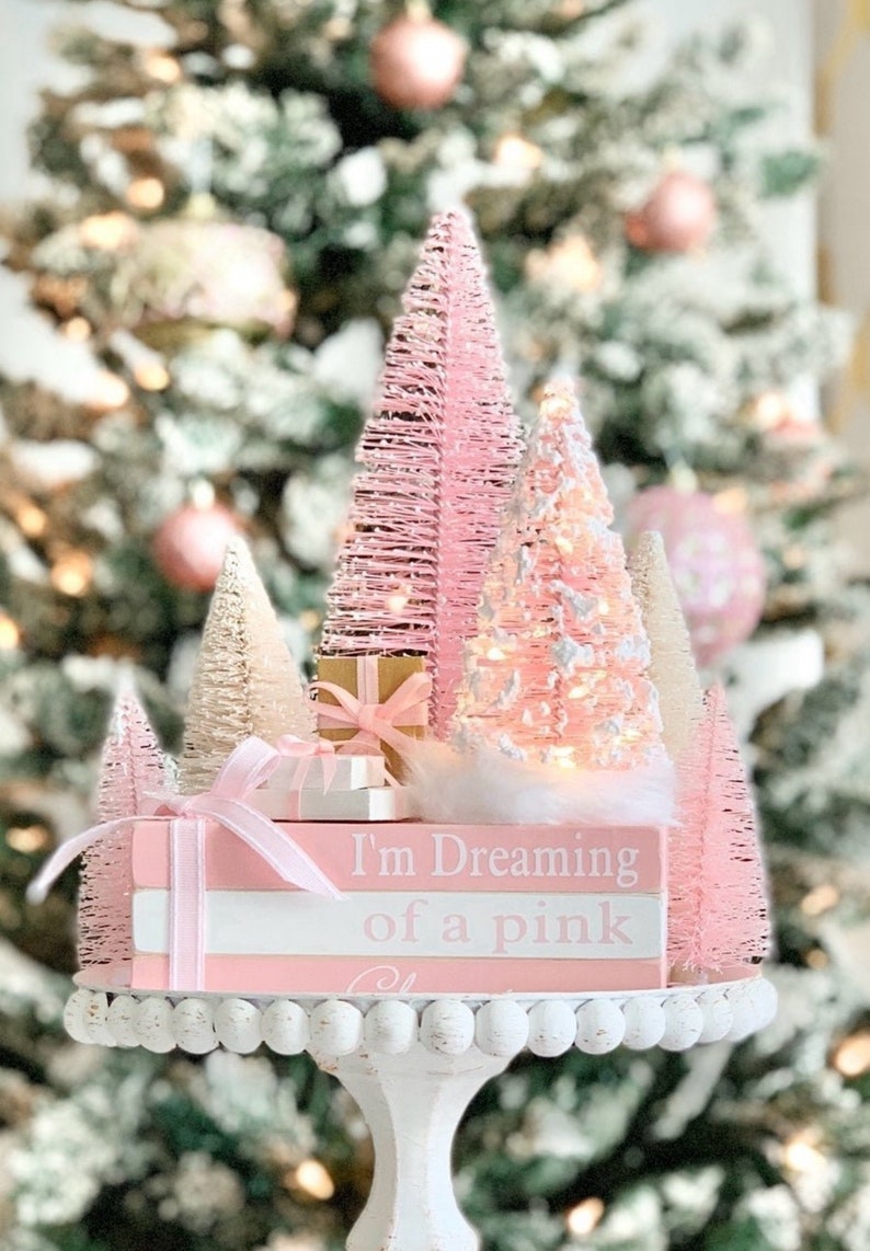 Puede incluir: Un conjunto de mini &aacute;rboles de Navidad rosas y blancos con luces, una caja de regalo rosa y una pila de libros rosas con el texto "I'm Dreaming of a Pink Christmas". Los &aacute;rboles est&aacute;n sobre un pedestal blanco con un borde de cuentas.