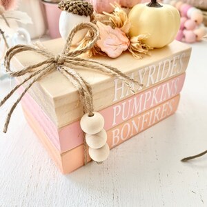 Puede incluir: Una pila de tres libros de madera con bordes pintados de rosa y naranja. Los libros est&aacute;n decorados con un lazo de arpillera, una peque&ntilde;a bellota, una hoja rosa y una calabaza amarilla. Los libros est&aacute;n etiquetados como "Hayrides", "Pumpkins" y "Bonfires".