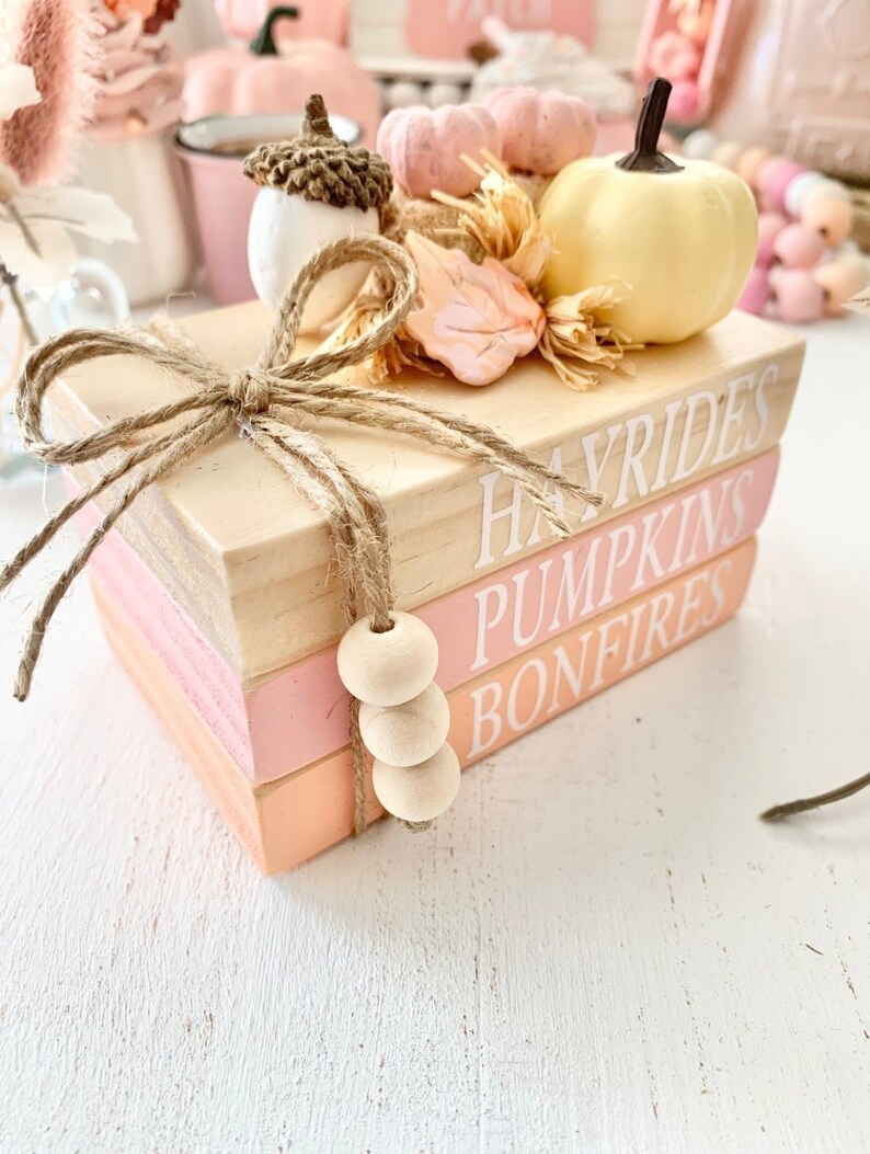 Puede incluir: Una pila de tres libros de madera con bordes pintados de rosa y naranja. Los libros est&aacute;n decorados con un lazo de arpillera, una peque&ntilde;a bellota, una hoja rosa y una calabaza amarilla. Los libros est&aacute;n etiquetados como "Hayrides", "Pumpkins" y "Bonfires".