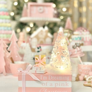 Puede incluir: Una pila de libros rosas con una cinta blanca y un peque&ntilde;o &aacute;rbol de Navidad rosa con luces blancas. Los libros est&aacute;n decorados con el texto "I'm Dreaming of a Pink Christmas".