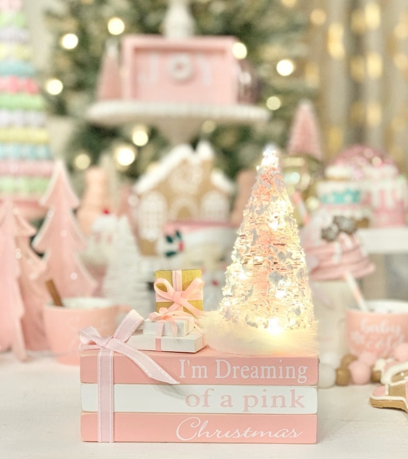 Puede incluir: Una pila de libros rosas con una cinta blanca y un peque&ntilde;o &aacute;rbol de Navidad rosa con luces blancas. Los libros est&aacute;n decorados con el texto "I'm Dreaming of a Pink Christmas".
