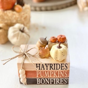 May include: A stack of three wooden blocks with the words "Hayrides", "Pumpkins", and "Bonfires" painted on them. The blocks are decorated with small pumpkins, acorns, and a fall leaf.