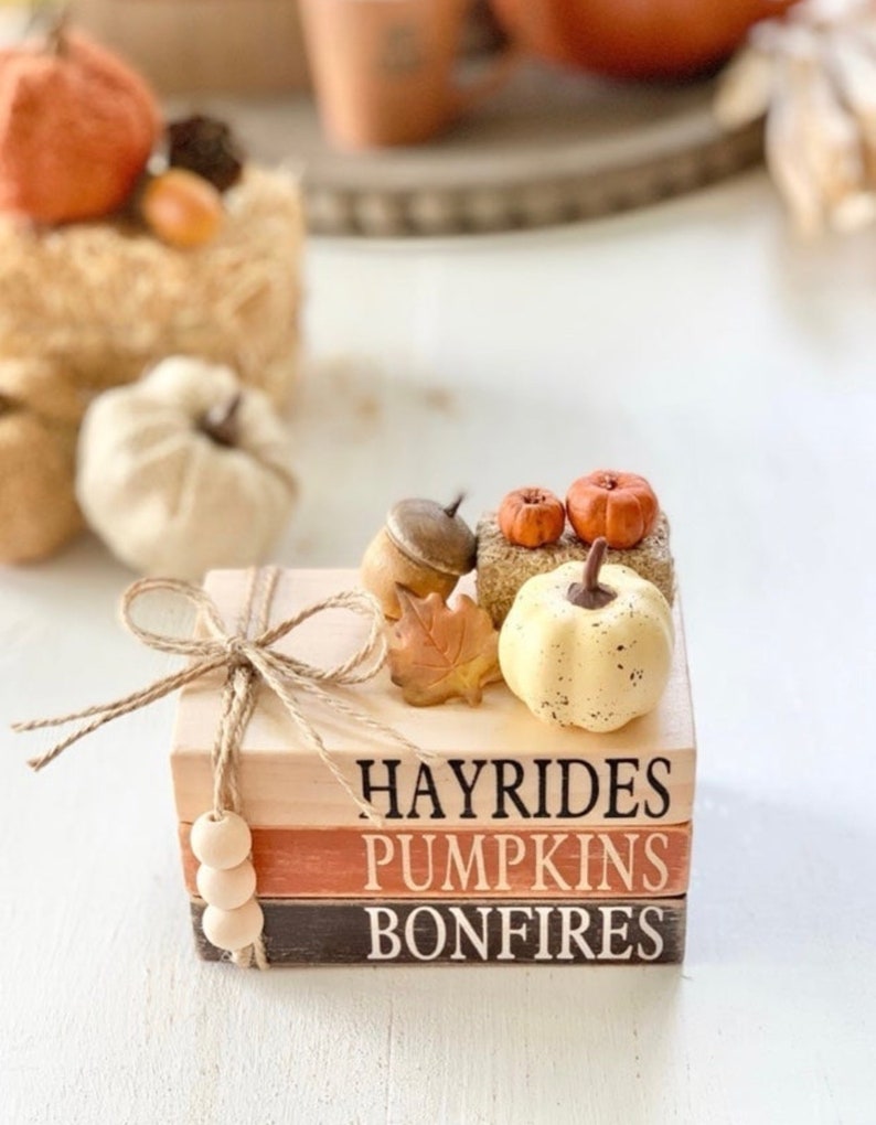 May include: A stack of three wooden blocks with the words "Hayrides", "Pumpkins", and "Bonfires" painted on them. The blocks are decorated with small pumpkins, acorns, and a fall leaf.