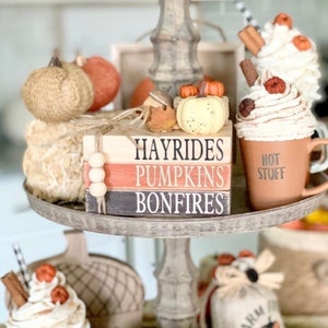 May include: A tiered tray decorated with fall-themed items, including pumpkins, a mug with whipped cream and cinnamon sticks, and a stack of wooden blocks with the words "Hayrides, Pumpkins, Bonfires".
