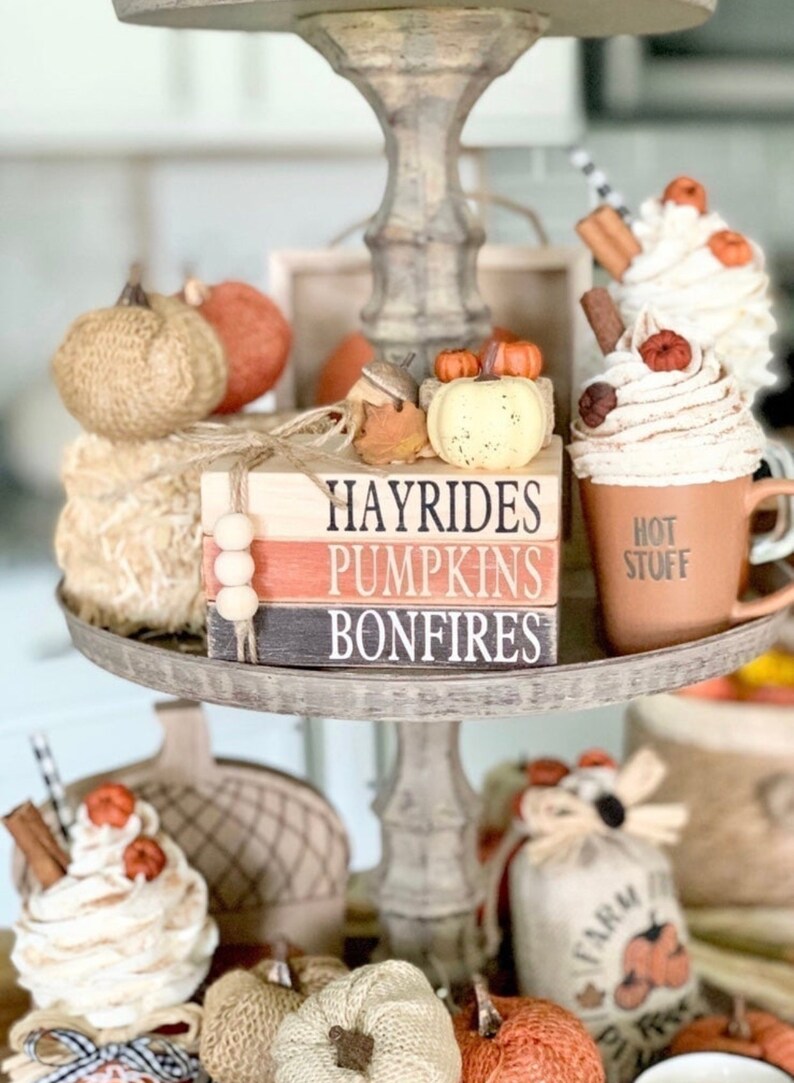 May include: A tiered tray decorated with fall-themed items, including pumpkins, a mug with whipped cream and cinnamon sticks, and a stack of wooden blocks with the words "Hayrides, Pumpkins, Bonfires".