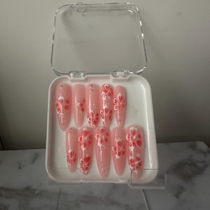 May include: A set of ten long, almond-shaped artificial nails in a clear plastic case. Each nail is painted a pale pink with a floral design in red and white. Some nails have small silver embellishments.
