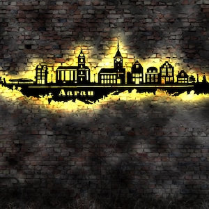 May include: A black metal silhouette of the city skyline of Aarau, Switzerland, with the city name below. The silhouette is illuminated with a warm yellow light and is mounted on a brick wall.