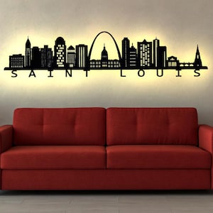 Könnte beinhalten: Schwarze Metall-Silhouette der Skyline von Saint Louis mit beleuchteten Gebäuden und dem Stadtnamen darunter. Das Kunstwerk hängt über einem roten Sofa, flankiert von Topfpflanzen, wodurch ein modernes Innendesign entsteht.