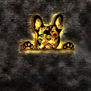 May include: A black silhouette of a French bulldog peeking over a brick wall. The dog's head and paws are illuminated with a warm yellow glow.