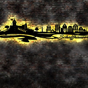 May include: A black metal wall art silhouette of the Schaffhausen skyline, illuminated with a warm yellow glow against a brick wall. The city's name is written in cursive below the skyline.